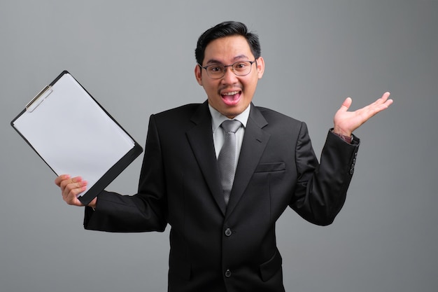 Excited handsome young businessman in formal suit holds a clipboard with papers document and shrugging hand isolated on grey background