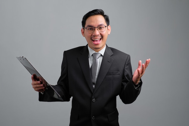 Excited handsome young businessman in formal suit holds a clipboard with papers document and shrugging hand isolated on grey background
