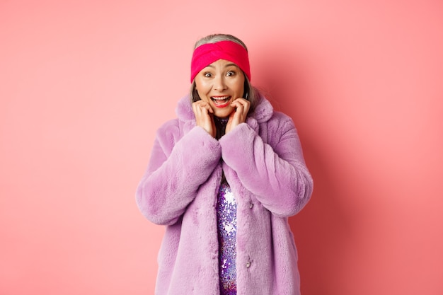 Excited and happy asian female senior model looking at camera with amazement, checking out promo offer, standing in trendy clothes over pink background.