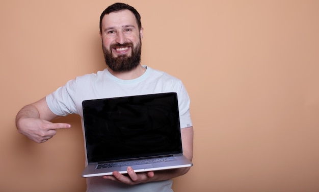 興奮した幸せなひげの男が空の画面のラップトップコンピューターを指さしている
