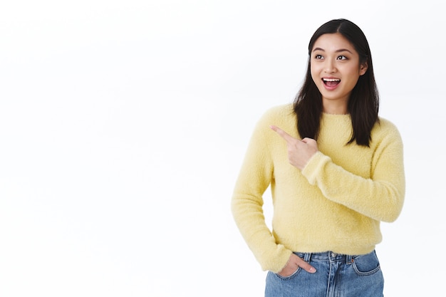 Eccitata felice bella studentessa asiatica in maglione giallo, sorridente e ansimante stupita, vedendo un prodotto meraviglioso, promozione fantastica, puntando il dito a sinistra nello spazio bianco vuoto della copia