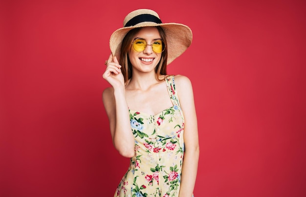 선글라스, 드레스, 여름 모자를 쓴 흥분되고 행복한 아름다운 젊은 여성은 분홍색 배경에서 포즈를 취하는 동안 재미있습니다.