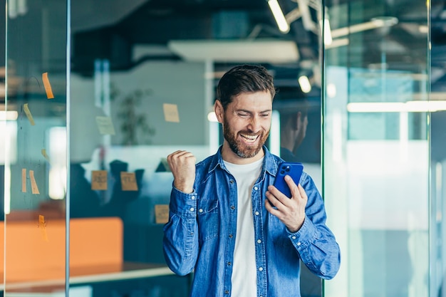 Eccitato uomo d'affari felice con la barba un vincitore professionista che celebra il successo leggendo buone notizie su uno smartphone entusiasta della messaggistica mobile