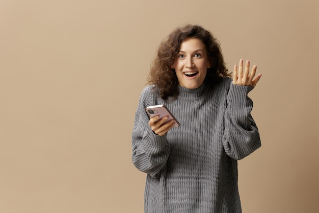 Excited happy curly beautiful lady in gray casual sweater get cool news from message posing isolated on over beige pastel background Social media network distance communication concept Copy space