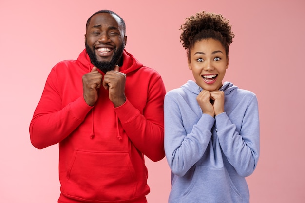 Excited happy girlfriend boyfriend smiling broadly nervously clenching fists pressed chest looking camera thrilled anticipate good news trying luck playing lottery together, standing pink background