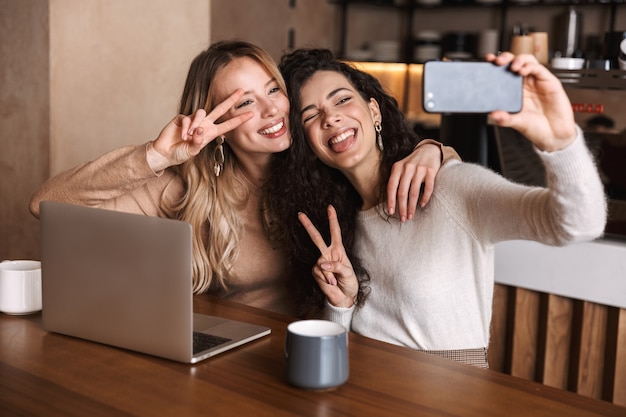 ラップトップコンピューターを使用してカフェに座っている興奮した幸せな女の子の友人は電話で自分撮りを取ります