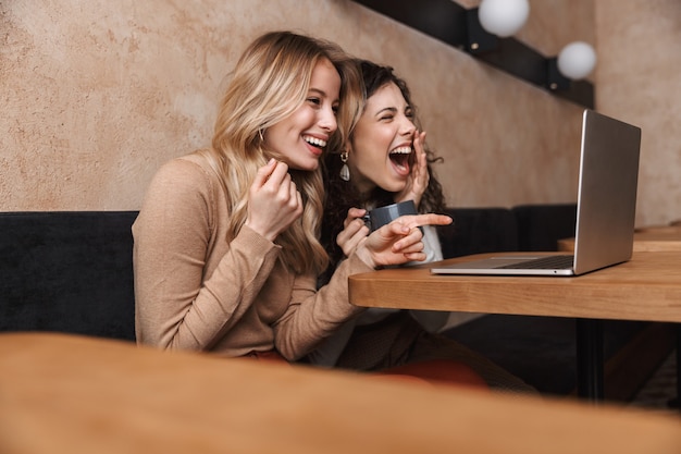 excited happy laughing pretty girls friends sitting in cafe using laptop computer pointing