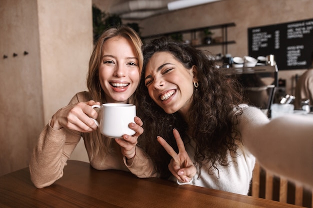 excited happy pretty girls friends sitting in cafe take a selfie by camera