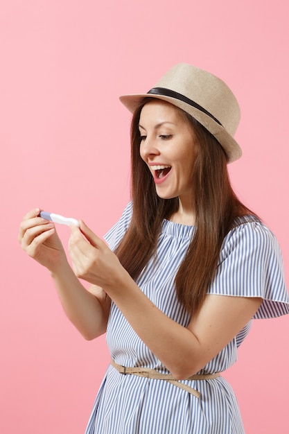 Excited happy woman in blue dress, hat hold in hand, looking at pregnancy test isolated on pink background. Medical healthcare gynecological, pregnancy fertility maternity people concept. Copy space.