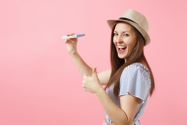 Excited happy woman in blue dress, hat hold in hand, looking at pregnancy test isolated on pink background. Medical healthcare gynecological, pregnancy fertility maternity people concept. Copy space.