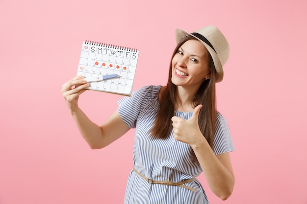 Excited happy woman in blue dress, hat hold in hand pregnancy test, periods calendar for checking menstruation days isolated on pink background. Medical, healthcare, gynecological concept. Copy space.
