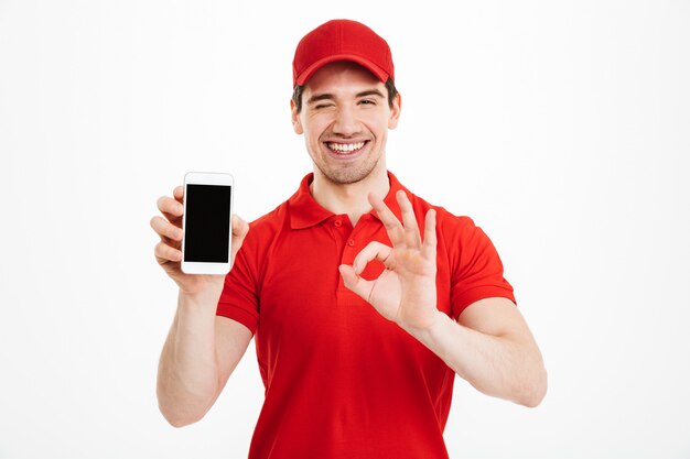 Excited happy young delivery man showing display of mobile phone make okay gesture.