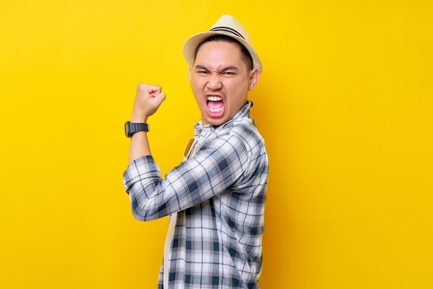 Excited happy Young handsome ethnic Asian man 20s wearing casual clothes hat showing biceps muscles on his hand demonstrating strength and power isolated on yellow background People lifestyle concept
