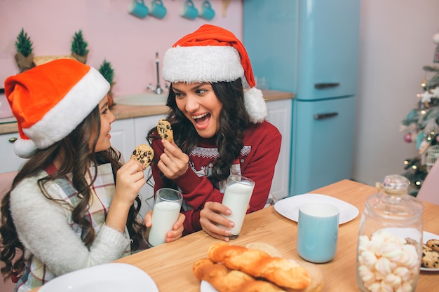 Excited and happy young woman and girl look at each other and hold cookies in hands. There are glasses of milk in their hands. People wear read hats. They sit in kitchen.