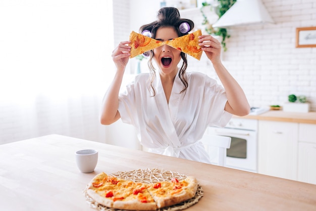 Excited happy young woman in kitchen plaing with pizza slices. Cover eyes with food. Carefree housekeeper very playful. Wear white dressing gown. Careless houseswife.