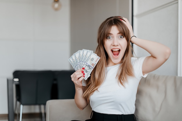 Excited happy young woman with money dollar bills in hand screaming sitting on a couch at home