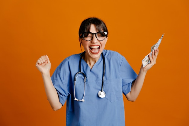 Excited holding cash young female doctor wearing uniform fith stethoscope isolated on orange background