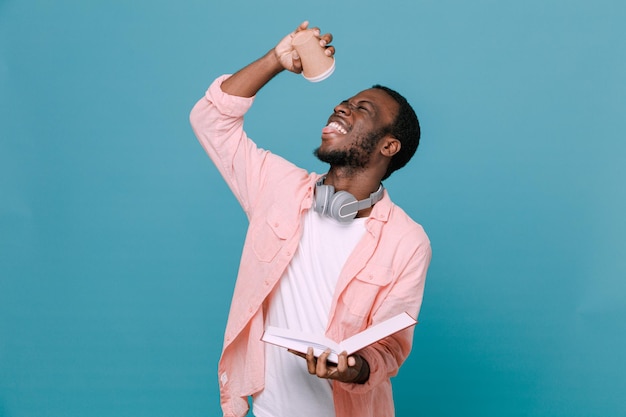 Eccitato tenendo la tazza di caffè con il libro giovane ragazzo afroamericano che indossa le cuffie sul collo isolato su sfondo blu