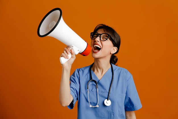 Excited holding loudspeaker young female doctor wearing uniform fith stethoscope isolated on orange background