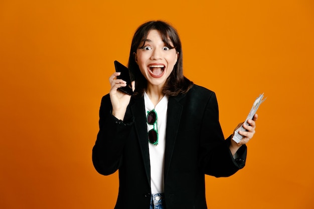 Excited holding money and phone young beautiful female wearing black jacket isolated on orange background