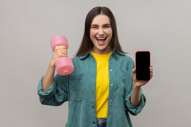 Excited holding pink dumbbell and blank screen smartphone in hands satisfied with sports app