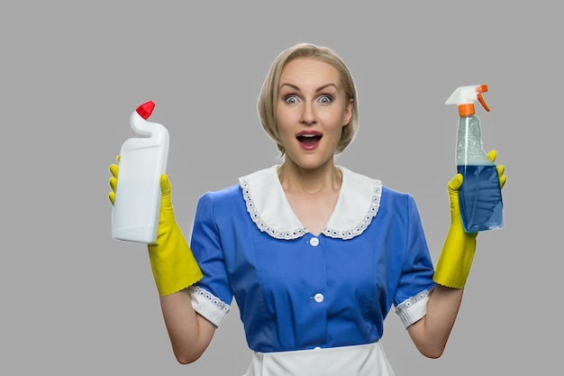 Excited housekeeping woman presenting cleaning supplies. Portrait of happy housekeeper holding cleaning equipment against gray background. Cleaning service discount.