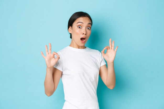 Excited and impressed asian girl in white t-shirt look surprised and astonished with amazing, perfect service, showing okay gesture and looked astounded over blue wall