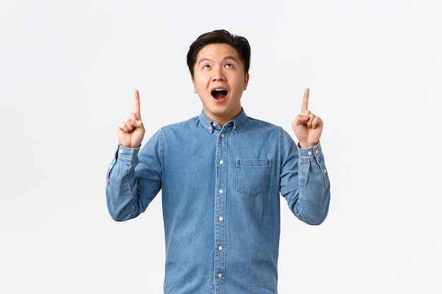 Excited and impressed asian young man open mouth, saying wow fascinated, checking out promo, looking and pointing fingers up at advertisement, stare at awesome banner, white background.