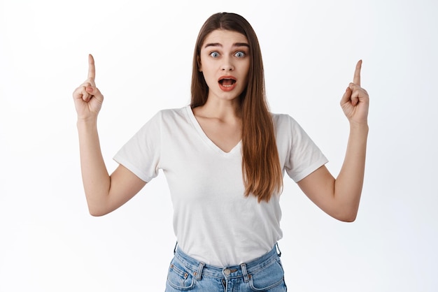 Excited and impressed caucasian woman pointing fingers up showing advertisement with dropped jaw staring astonished at camera check this out gesture white background