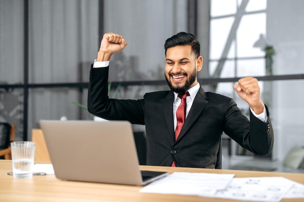 Photo excited indian or arabian businessman in suit sits in a modern office looking at laptop happy by good news rejoice by win raising hand in yes gesture celebrating project success big profit