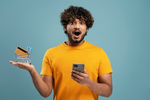 Excited Indian man using mobile phone, holding shopping trolley with credit card. Shopping, sales