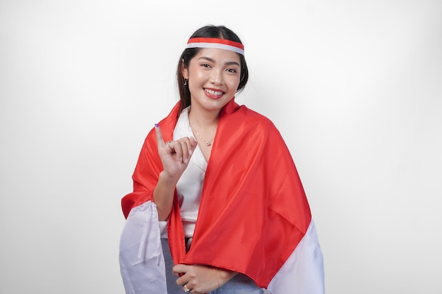 Excited Indonesian woman proudly showing little finger dipped in purple ink after voting for president and parliament election wearing mini flag headband and white shirt covering flag on the back