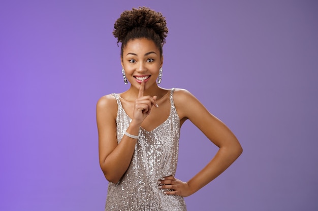 Excited joyful african-american young 20s woman in silver dress telling secret gossiping showing shh gesture say shush smiling gladly wanna make surprise, hold hand waist index finger on mouth.