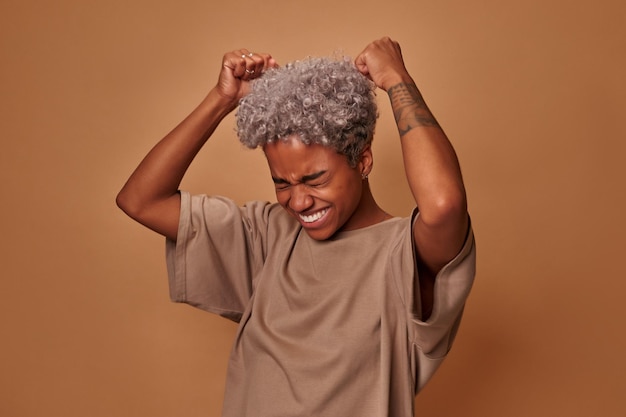 Excited joyous young cute african american woman raises clenched fists stands without make up agains...