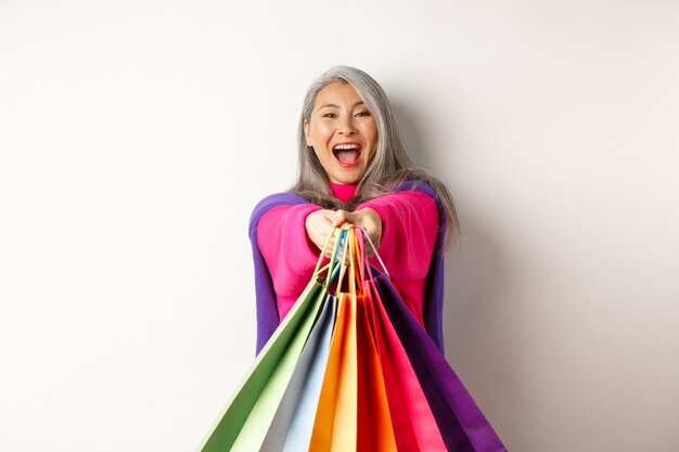 Photo excited korean lady with grey hair, laughing and showing shopping bags, visit stores with special discounts, standing over white background.