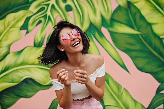 Photo excited lady in pink glasses laughing at camera outdoor shot of charming girl posing near graffiti