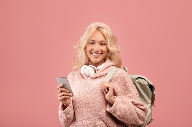 Excited lady student using smartphone and smiling at camera while standing with backpack over pink