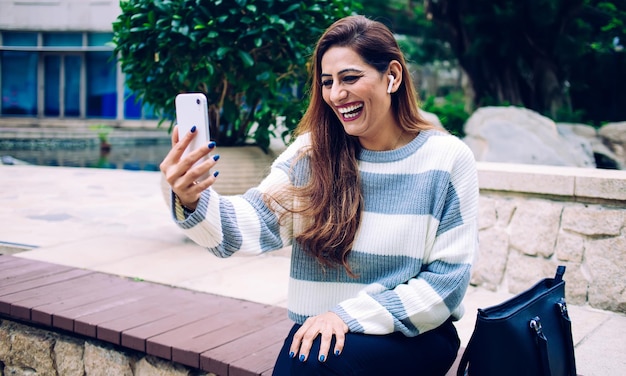 Excited laughing adult woman with earbud in casual striped sweater streaming on smartphone