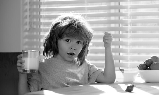 Excited little boy holding glass of milk sitting at wooden table in kitchen adorable child kid enjoy