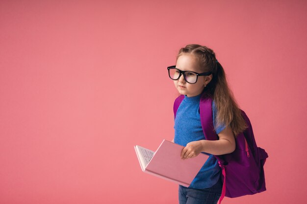 Excited little girl in glasses with school bag and holding book goes to school