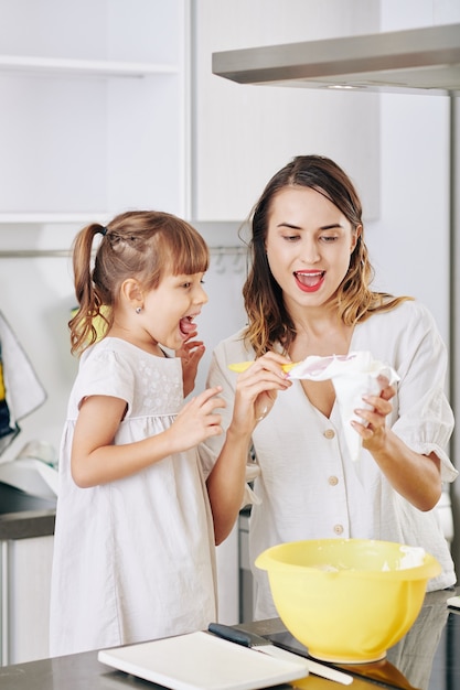 Bambina emozionante guardando sua madre che riempie la sacca da pasticcere con panna montata quando si prepara la torta di compleanno a casa