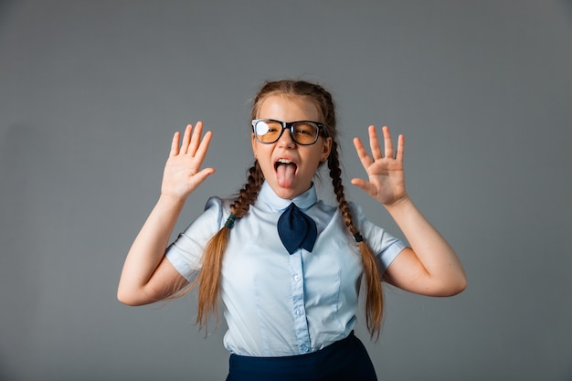 Excited little girl in school uniform and glasses having fun and showing ten fingers while standing against gray background