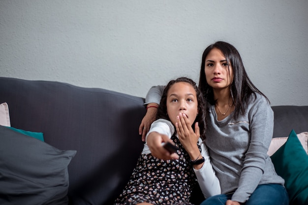 Bambina emozionante che guarda la tv con sua madre sul divano di casa con spazio vuoto per il testo.