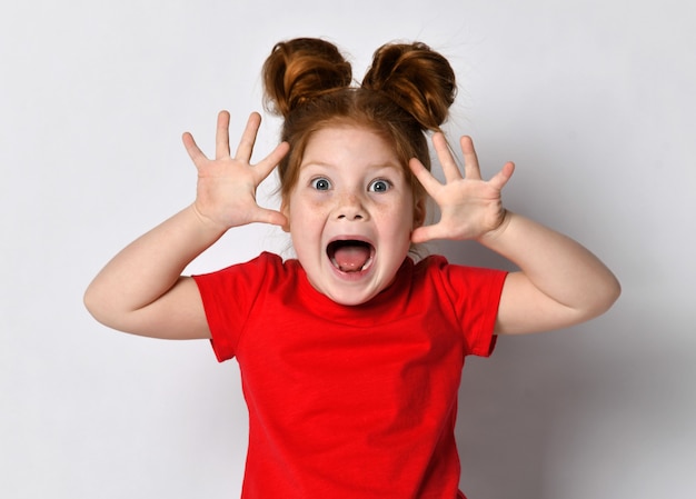 Excited Little girl with freckles and red hair screams loudly with raised arms, looking at the camera. Portrait of emotional child on gray background. The concept of childrens hysterics and emotions.