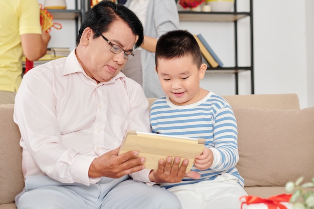 Excited little Vietnamese boy and his grandfather watching cartoon on tablet computer