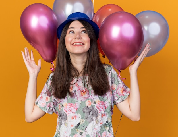 Excited looking up young beautiful girl wearing party hat standing in front balloons spreading hands 