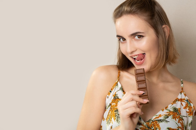 Excited lovely brunette woman holding a small chocolate bar on a grey studio background. Empty space