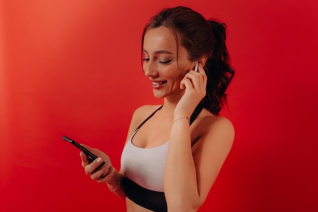 Excited lovely girl with wavy dark hair is wearing sport top is listening music in headphones and holding smartphone before training