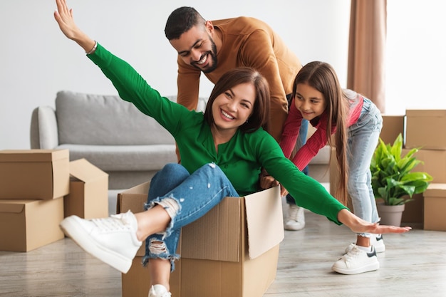 Excited loving family having fun celebrating moving day, cheerful Arab husband and daughter playing together, riding mom in cardboard box container in living room. New Home And Real Estate Concept