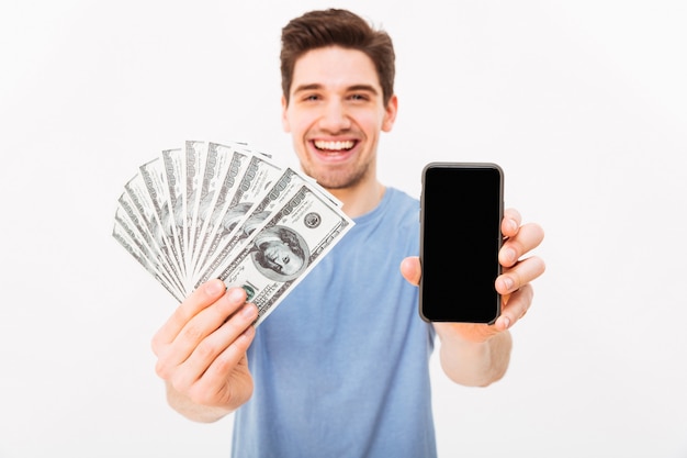 Excited man in casual t-shirt demonstrating his money prize and copyspace screen of smartphone, isolated over white wall